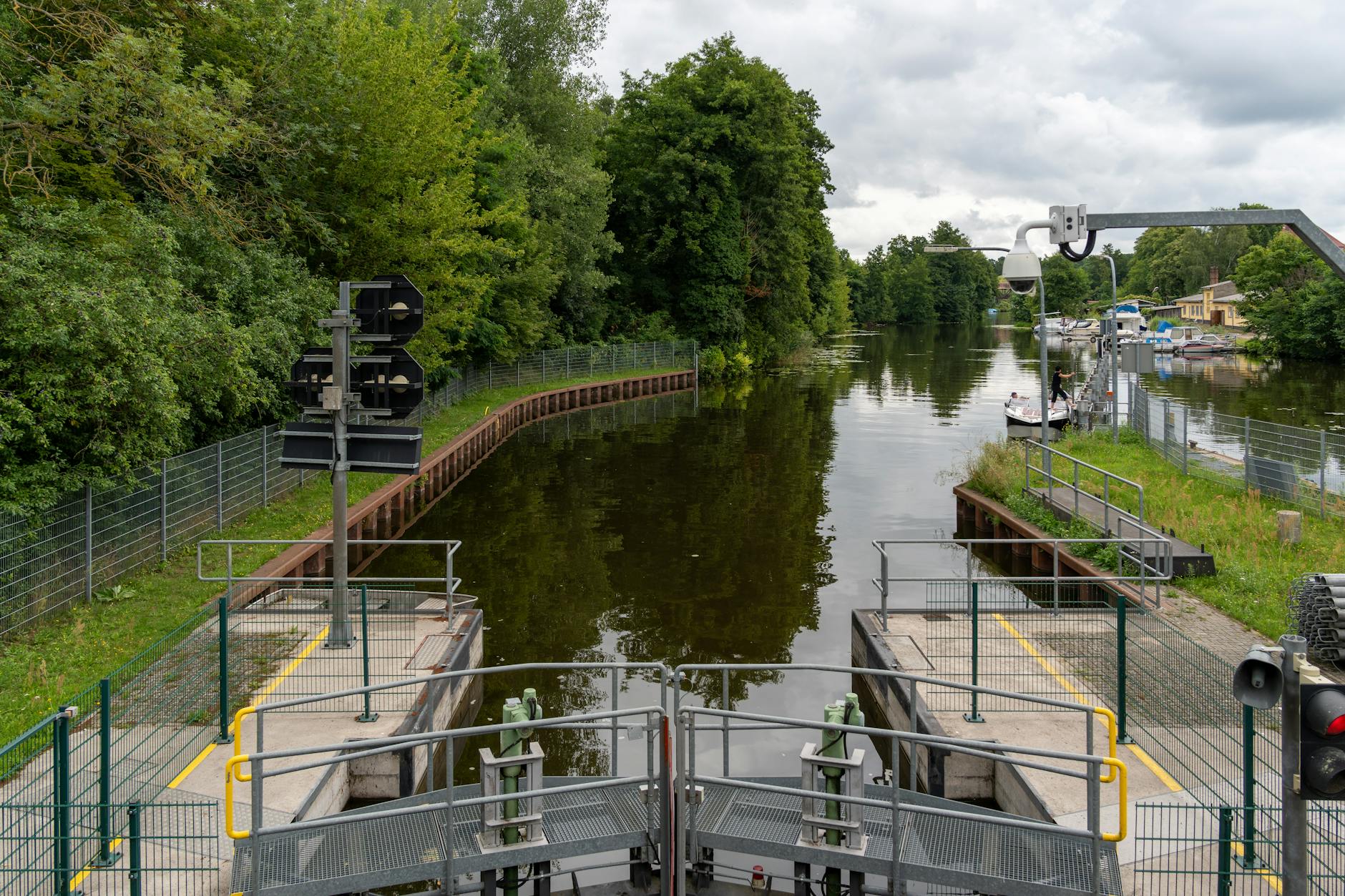 Peaceful canal scene featuring a water lock, lush greenery, and moored boats under a cloudy sky.