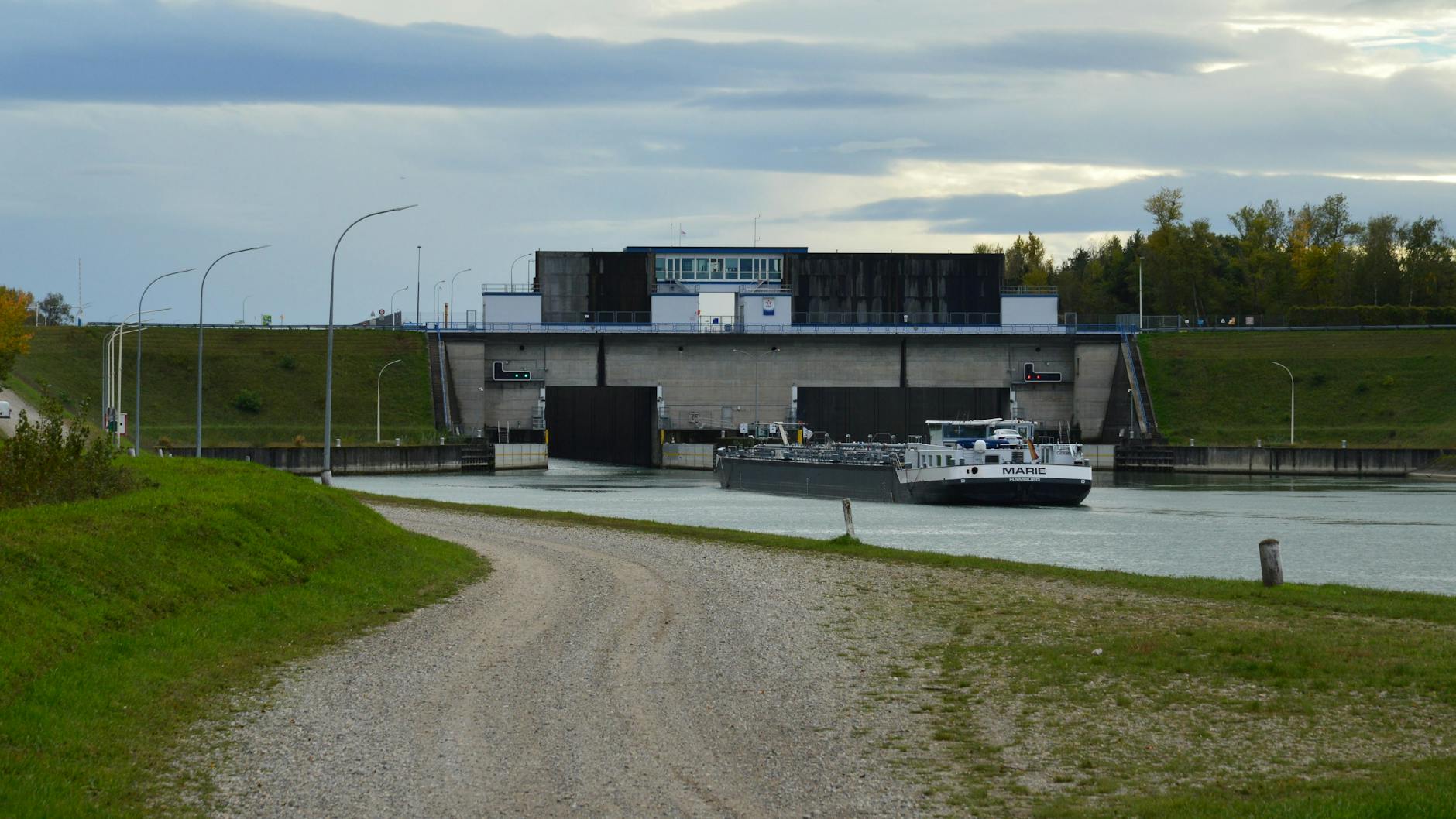 A cargo barge enters a lock on a river canal under an overcast sky.